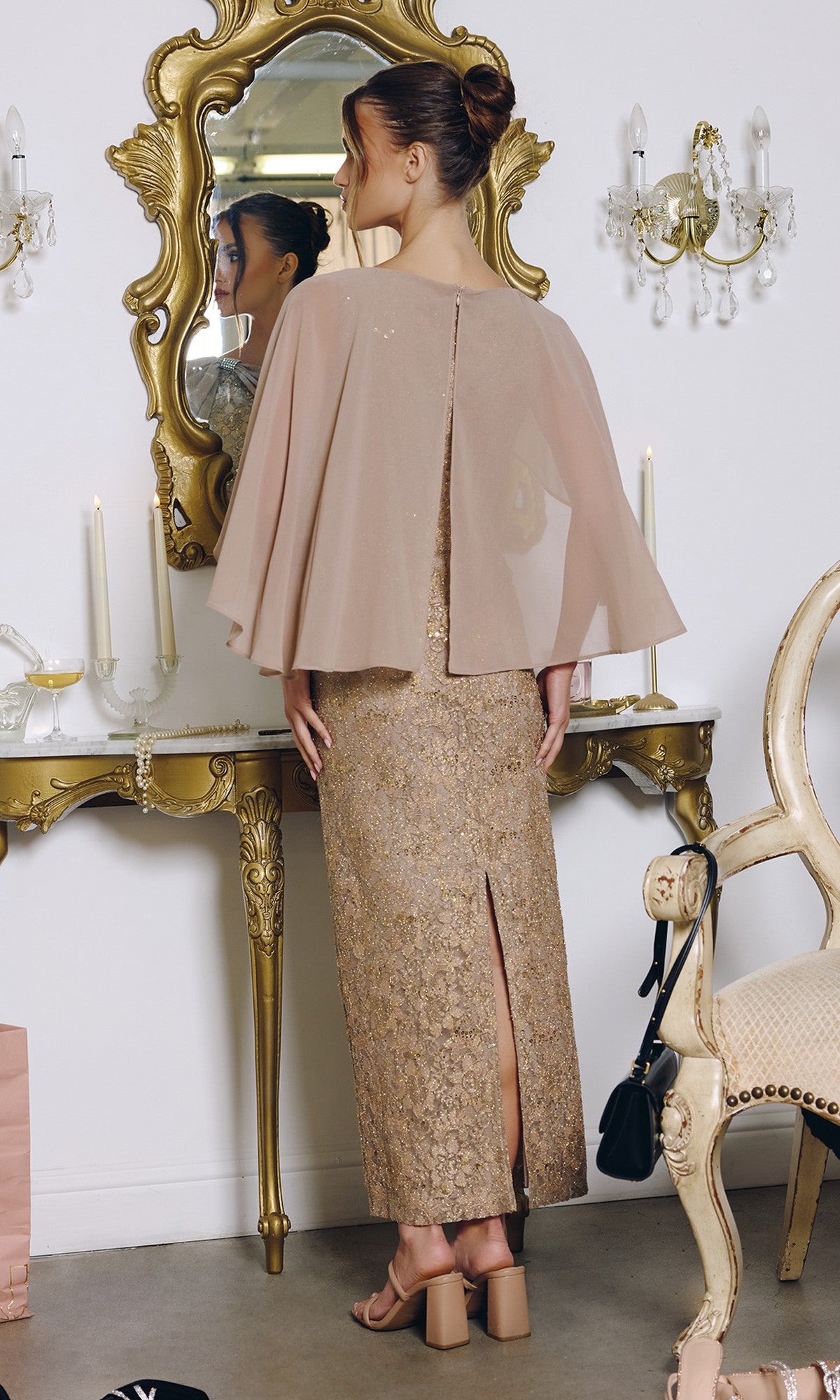 A woman in the Amoris T906 Long Prom Dress, featuring sequin lace and a sheer chiffon cape overlay, stands before a vintage vanity adorned with candles and ornate decor, admiring her reflection.